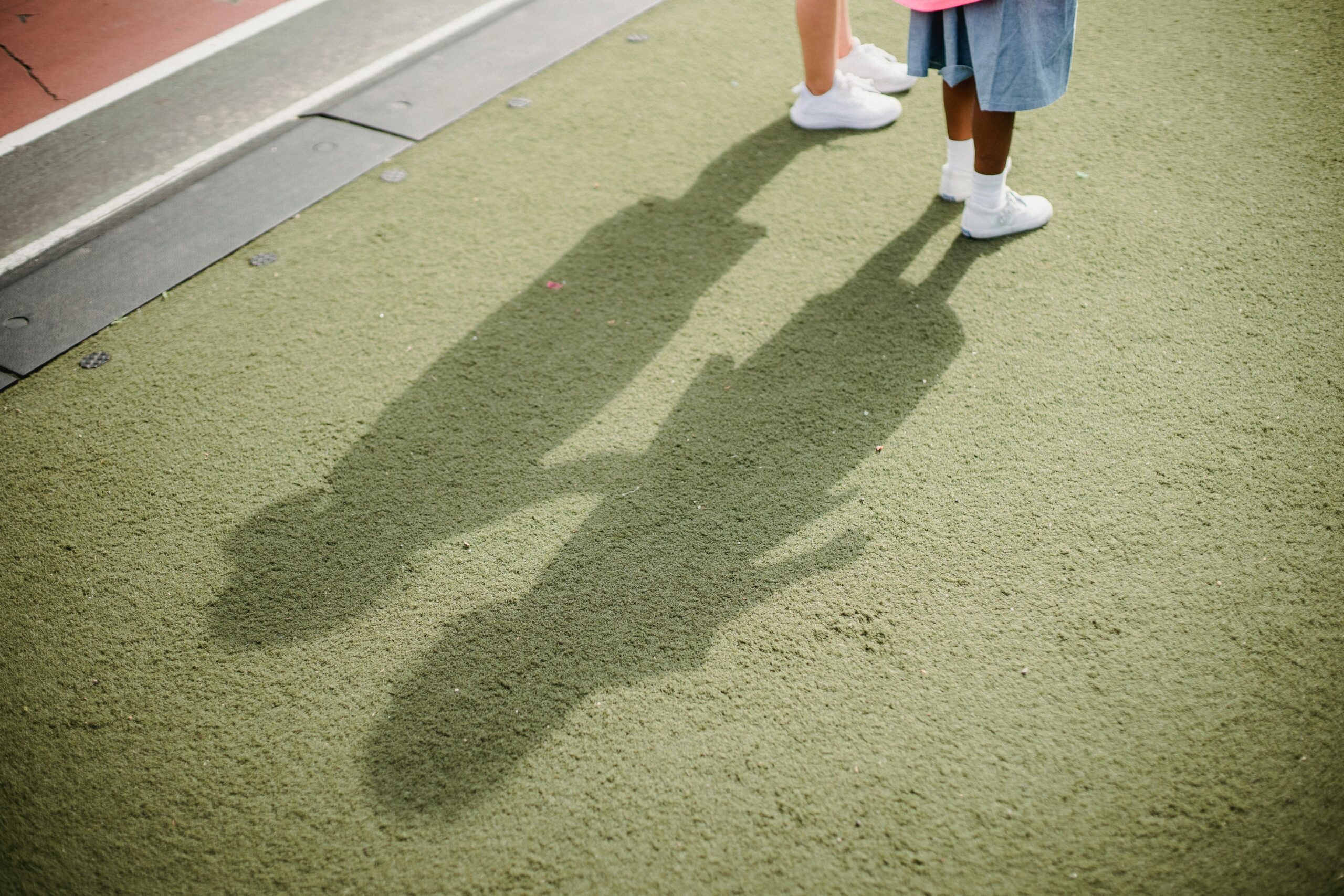 Two children casting shadows on a green school playground surface.