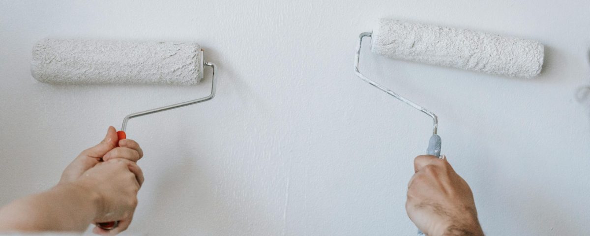 Close-up of two individuals painting a wall with paint rollers indoors.